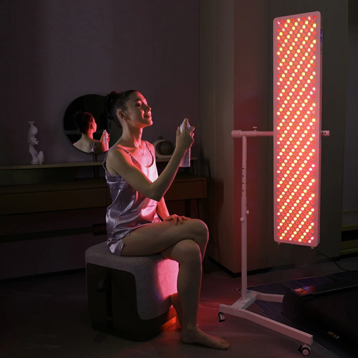 A woman seated beside a tall red light therapy panel in a dark room